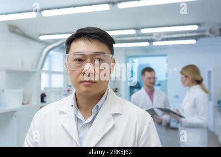 Portrait d'un jeune homme asiatique médecin scientifique debout dans un laboratoire d'un centre de recherche dans un manteau blanc et des lunettes de protection et regardant sérieusement la caméra. Les collègues travaillent ensemble. Banque D'Images