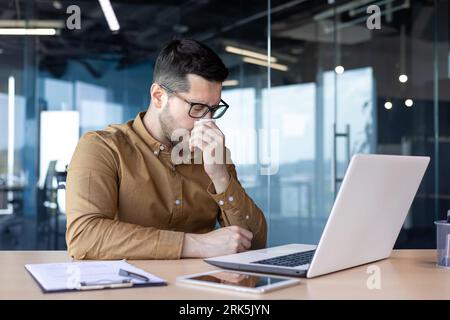 Un jeune employé de bureau se sent malade au travail. Il est assis à une table avec un ordinateur portable et s'essuie le nez avec une serviette. souffrant du rhume. Banque D'Images