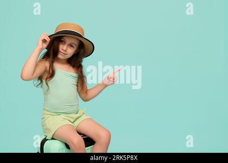 Adorable petite fille en tenue d'été tendance et chapeau de paille, pointant de côté un espace de copie ad sur fond bleu isolé, assis sur une valise, smi Banque D'Images