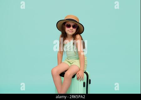 Belle petite fille en tenue d'été, chapeau de paille et lunettes de soleil, assis sur une valise, souriant regardant la caméra, isolé sur fond bleu. Banque D'Images