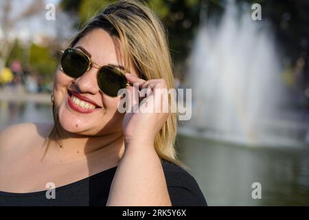 gros plan portrait de jeune femme blonde plus taille latina, debout dans le parc public heureux portant des lunettes de soleil, posant souriant et regardant la caméra, avec Banque D'Images