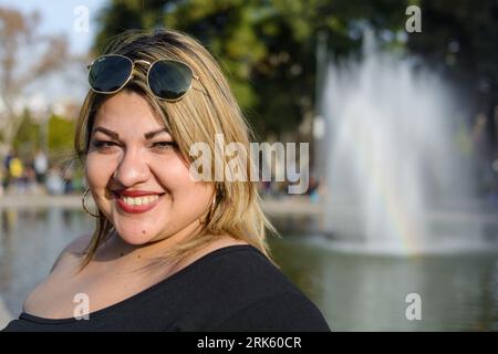 gros plan portrait de jeune femme blonde plus taille latina, debout dans le parc public heureux portant des lunettes de soleil, posant souriant et regardant la caméra, avec Banque D'Images