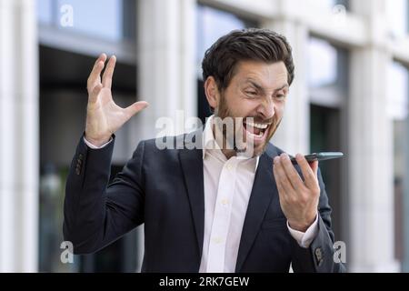 Jeune homme d'affaires furieux et indigné debout à l'extérieur des immeubles de bureaux parlant sur haut-parleur, criant fort et faisant des gestes avec les mains. Banque D'Images
