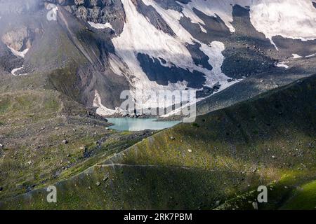 Un lac montagneux turquoise laiteux (peut-être l’un des lacs d’Abudelauri) se trouve au pied des champs de neige et des pentes de éboulis sur l’ascension vers le col de Chaukhi dans la municipalité de Dusheti, région de Mtskheta-Mtianeti, Géorgie. Des roches sombres traversent les champs de neige. De gros blocs de roche gisent sur la rive opposée. Une crête verte, partiellement ombragée, s’étend sur le premier plan. Un étroit sentier la traverse. Les trois lacs d’Abudelauri (géorgien აბუდელაურის ტბები), le lac Vert, le lac Bleu et le lac Blanc, se situent entre le col et le village de Roshka. Le col est à environ 3 340 mètres d’altitude et mène à Juta dans Kazbegi Mun Banque D'Images