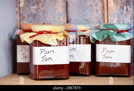 Pots fraîchement préparés de confiture de prunes maison, avec des étiquettes manuscrites et des toppers en tissu placés sur une étagère dans un placard en bois rustique Banque D'Images