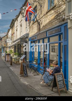 Une dame est assise et profite du soleil d'été avec un café devant un café sur la chaussée dans Black Jack Street, Cirencester, Gloucestershire. Banque D'Images