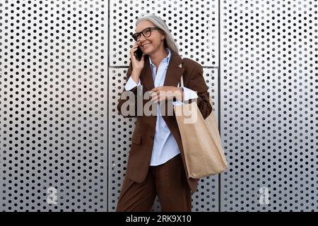 une femme d'âge moyen aux cheveux gris avec une coupe de cheveux bob vêtue d'une veste et d'un pantalon marche sur le fond d'un mur de bâtiment gris Banque D'Images