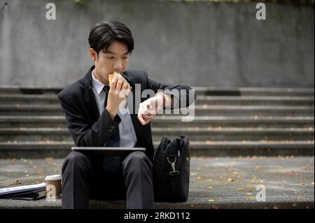 Un jeune homme d'affaires asiatique occupé et affamé ou un employé de bureau vérifie le temps sur sa montre et mange un sandwich pour son déjeuner rapide sur un escalier Banque D'Images