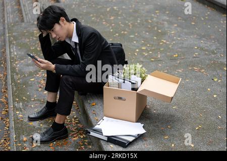 Malheureux et bouleversé jeune homme d'affaires asiatique ou employé de bureau masculin tapant sur son téléphone alors qu'il était assis sur un escalier avec une boîte de ses affaires personnelles afte Banque D'Images