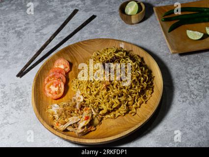 Mie goreng ayam geprek. Nouilles frites servies sur une assiette en bois accompagnées de poulet, tomates fraîches et saupoudrées d'oignons frits Banque D'Images