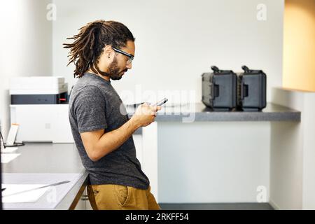 Vue latérale de l'homme d'affaires confiant utilisant le téléphone intelligent tout en se tenant par la table dans le bureau Banque D'Images