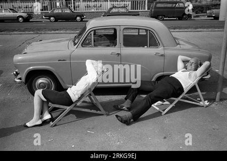 Couple des années 1970 au Seaside UK. Un couple d'âge moyen avec leur Austin Cambridge A50 et leurs propres chaises longues profitent d'un samedi après-midi endormi au soleil. Il s'est dépouillé de son gilet de corde et a décidé après tout de ne pas lire son livre, thats maintenant sur le terrain. Elle protège ses yeux du soleil. Southend on Sea, Essex, Angleterre 1974 HOMER SYKES Banque D'Images