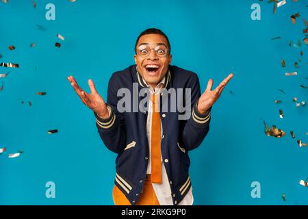 étudiant afro-américain joyeux et branché montrant le geste wow pendant la fête sous les confettis sur le bleu Banque D'Images