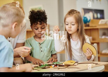 fille jouant avec du matériel didactique près d'enfants interraciaux et professeur à l'école montessori Banque D'Images