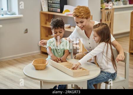 heureux professeur parlant à des enfants interraciaux jouant avec des matériaux didactiques en bois dans la classe montessori Banque D'Images