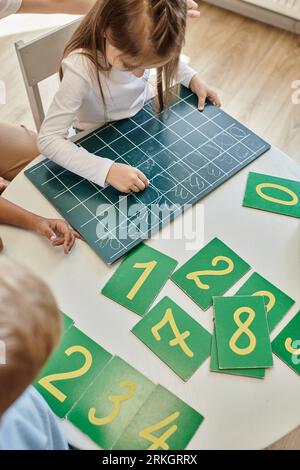 Vue de dessus d'une fille écrivant des numéros sur un tableau noir, apprenant à compter à l'école Montessori Banque D'Images