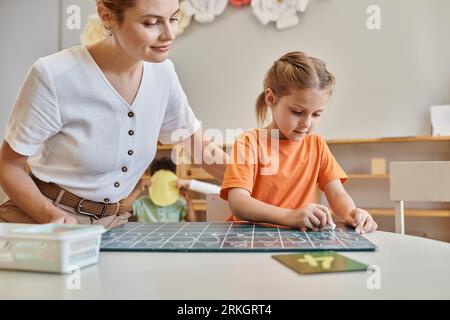 Professeur heureux observant fille intelligente dans l'école Montessori, comptant, écrivant au tableau noir, chiffres Banque D'Images