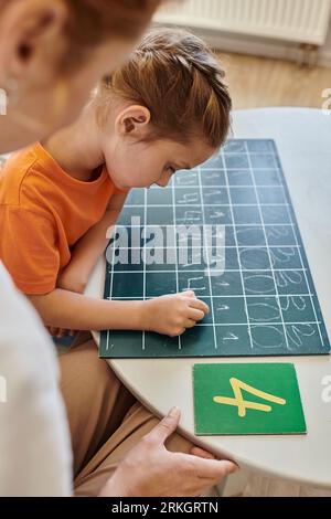 Enseignante observant la fille intelligente dans l'école Montessori, comptant, écrivant au tableau noir, les chiffres Banque D'Images