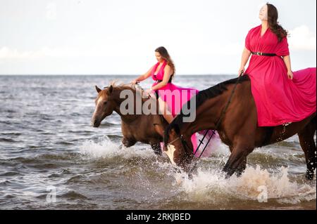 Les deux cavalières en robes roses vibrantes mènent deux chevaux majestueux le long d'une côte de plage. Banque D'Images