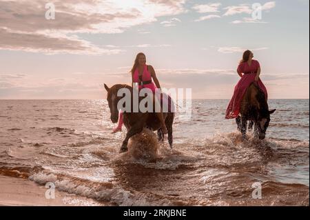 Les deux cavalières en robes roses vibrantes mènent deux chevaux majestueux le long d'une côte de plage. Banque D'Images