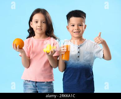 Petits enfants asiatiques avec du jus d'agrumes frais, orange et presse-étoupe montrant le geste du pouce vers le haut sur fond bleu Banque D'Images