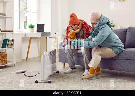 Couple congelé senior assis à la maison et essayant de réchauffer leurs mains sur le chauffage électrique. Banque D'Images