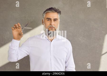 Portrait d'un coiffeur élégant brutal avec barbe tenant des ciseaux sur fond gris. Banque D'Images