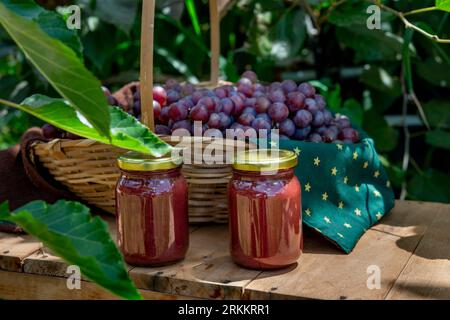 Confiture de raisin dans des bocaux en verre avec panier plein de raisins frais en arrière-plan sur une table en bois dans le jardin, bocaux sans étiquette Banque D'Images