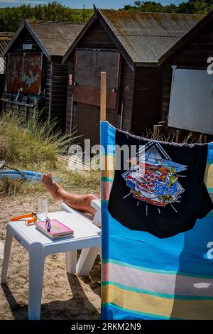 Résumé. Homme bronzer ses jambes et se détendre devant une cabane de plage typiquement britannique dans le Dorset. ROYAUME-UNI. Été. Ensoleillé. Aucun visage visible. Réservez et buvez. Banque D'Images