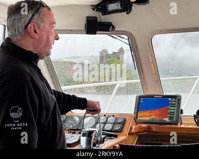 26 août 2023, Grande-Bretagne, Drumnadrochit : le skipper Ali Matheson se tient sur le pont d'un bateau sur le Loch Ness, les ruines du château d'Urquhart peuvent être vues en arrière-plan. En Écosse, samedi a commencé ce qui est considéré comme la plus grande recherche pour le monstre du Loch Ness, appelé Nessie, depuis des décennies. Des dizaines de volontaires du monde entier ont posté samedi matin sous une pluie battante partielle à 17 postes d'observation autour du célèbre lac dans les Highlands. Pendant la journée, des bateaux dotés d'une technologie spéciale comme un hydrophone - une sorte de microphone sous-marin - devaient naviguer sur le Loch Ness. L'utilisation du Dron Banque D'Images