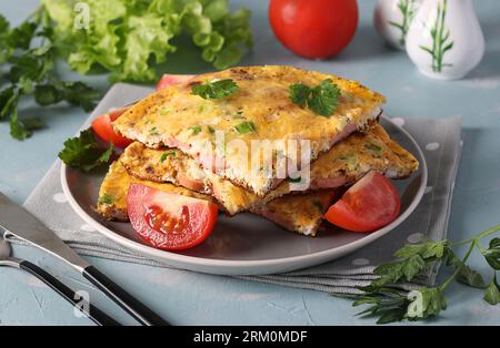 Omelette aux saucisses, fromage et persil servie avec des tomates sur une assiette grise sur fond bleu clair. Banque D'Images