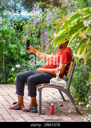 Homme noir assis sur un banc de parc avec le visage caché à l'aide d'un smartphone - Tours, Indre-et-Loire (37), France. Banque D'Images