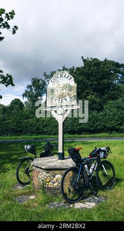 Deux vélos bikepacking sont garés contre un panneau de signalisation du village sur le green à Cockley Cley à Norfolk, Angleterre Royaume-Uni Banque D'Images