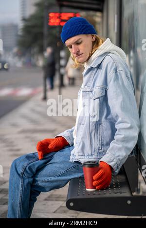 Jeune homme assis sur un banc à l'arrêt de bus. Guy avec tasse à café attendant calmement les transports en commun. Banque D'Images