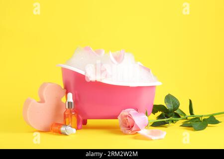 Petite baignoire avec mousse, éponge, bouteilles d'huile essentielle et fleur de rose sur fond jaune Banque D'Images