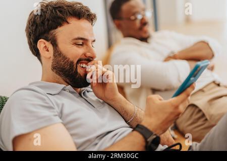 Employé masculin regardant quelque chose de drôle au téléphone et souriant tout en prenant une pause du travail au bureau, allongé sur un sac paresseux. Collègues divers r Banque D'Images