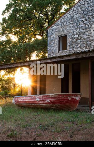 Soleil brillant à travers les arbres sur le petit bateau en bois rouge devant une maison en pierre à Umag, Croatie. Banque D'Images