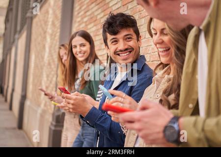 Vue latérale multiracial groupe souriant jeune génération z penchant mur de briques à l'aide de téléphones à l'extérieur. Banque D'Images