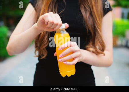 Jeune femme méconnaissable ouvre une bouteille de limonade orange en plastique sur un jour d'été à l'extérieur, debout dans une rue de la ville contre dans des bâtiments colorés Banque D'Images