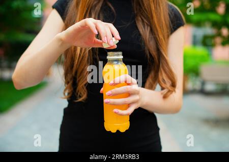 Jeune femme méconnaissable ouvre une bouteille de limonade orange en plastique sur un jour d'été à l'extérieur, debout dans une rue de la ville contre dans des bâtiments colorés Banque D'Images