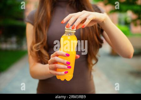 Jeune femme méconnaissable ouvre une bouteille de limonade orange en plastique sur un jour d'été à l'extérieur, debout dans une rue de la ville contre dans des bâtiments colorés Banque D'Images