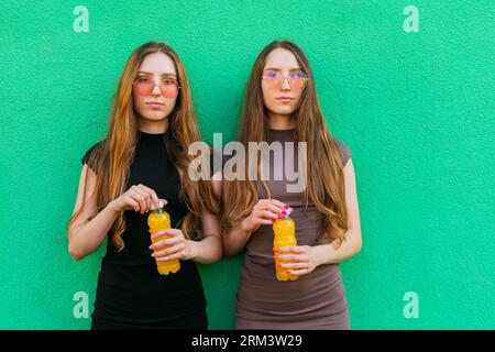 Jeunes sœurs jumelles mignonnes avec des lunettes de soleil cool tenant des bouteilles en plastique limonade jaune debout sur un fond de mur vert Banque D'Images