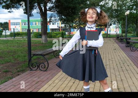 Fille avec sac à dos, uniforme scolaire avec des arcs blancs et pile de livres près de l'école. Retour à l'école, élève heureux, manuels lourds. Éducation, primaire sc Banque D'Images