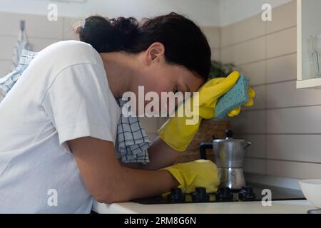 Une femme au foyer, fatiguée, frotte le poêle avec des gants. Le travail de routine domestique des femmes Banque D'Images