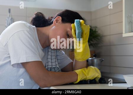 Une femme au foyer, fatiguée, frotte le poêle avec des gants. Le travail de routine domestique des femmes Banque D'Images