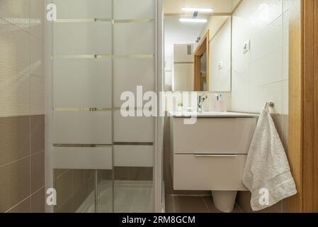 Salle de bains avec petit lavabo en porcelaine blanche avec armoire à tiroirs en dessous, miroir sans cadre et cabine de douche carrée simple Banque D'Images
