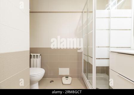 Salle de bains avec un petit évier en porcelaine blanche avec une unité de tiroir ci-dessous, un miroir sans cadre, une échelle de sol, des carreaux blancs et bruns, et un simple sho carré Banque D'Images