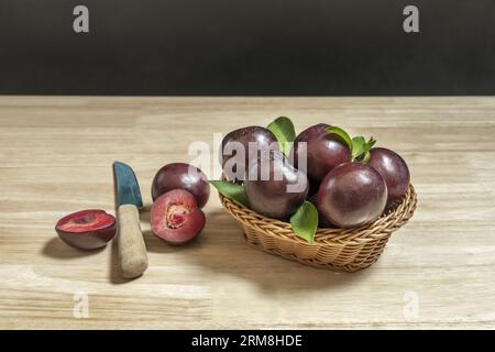 Un panier plein de prunes rouges mûres avec une moitié coupée à côté d'un couteau sur une table en bois Banque D'Images