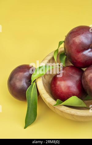 Un bol en bois de bambou rempli de prunes rouges mûres sur une surface jaune lisse Banque D'Images