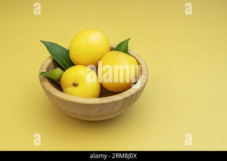 Un petit bol en bois de bambou rempli de grandes prunes jaunes mûres avec des feuilles vertes sur une surface jaune complètement lisse Banque D'Images